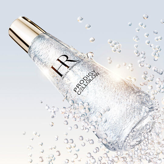 Flacon de serum Helena Rubinstein Prodigy Cellglow rempli de bulles, incline et entoure de bulles eclatantes en suspension, sur un fond clair et lumineux.