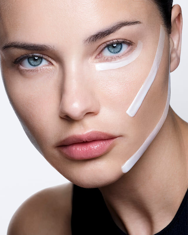 Adriana Lima mit weißer Creme und auf das Gesicht aufgetragenen Streifen, die einen sichtbaren Lifting-Effekt zeigen.