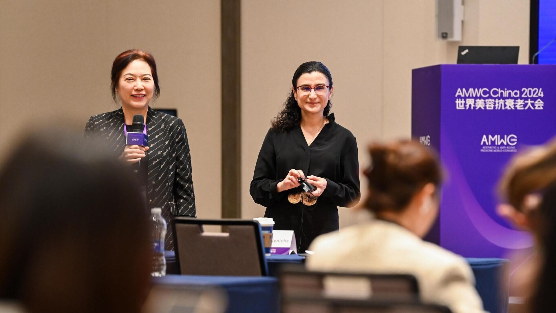 Two speakers, one in a black blouse and the other in glasses, present at the AMWC China 2024 conference.