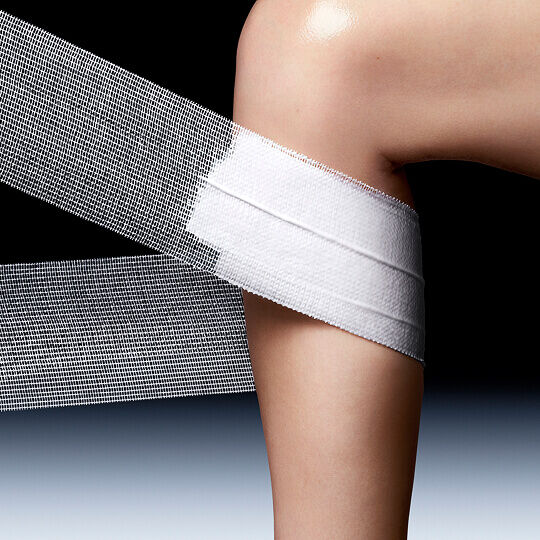 Ein Knie, das teilweise mit einem breiten, strukturierten weißen Netzband bedeckt ist, vor dunklem Hintergrund, das an Fursorge oder einen Verband erinnert.