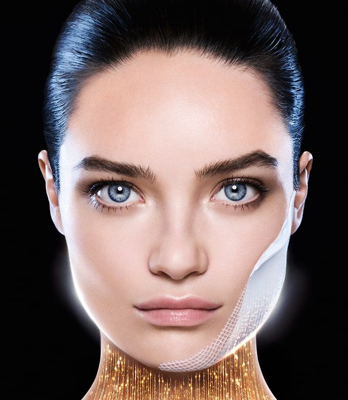 Frauengesicht mit blauen Augen, ein feines Patch auf der Wange. Unter dem Kinn symbolisiert ein goldener Glanz einen straffenden oder revitalisierenden Effekt. Schwarzer Hintergrund.