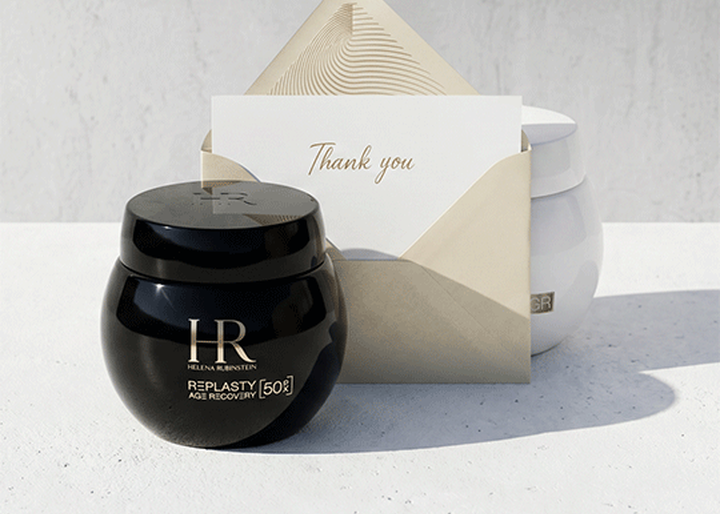 Un pot noir de creme Helena Rubinstein Replasty Age Recovery et une enveloppe contenant une carte "Thank you" poses sur une surface blanche, avec un pot blanc en arriere-plan.